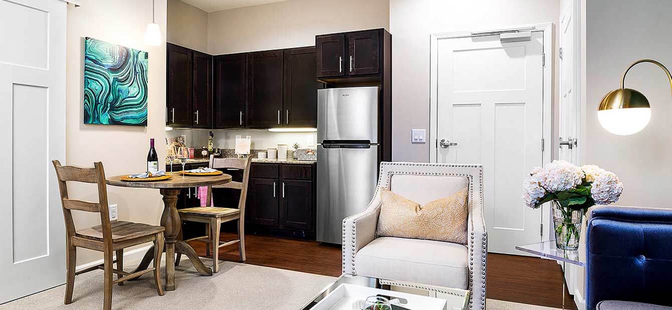 Image displays interior of a Heartis suite. Kitchenette is in the background with cabinets and stainless steel refrigerator. A small dining area is in front of the kitchenette. A sliding barn door opening onto another room is at the left side of the image. On the right of the image is large tufted arm chair.