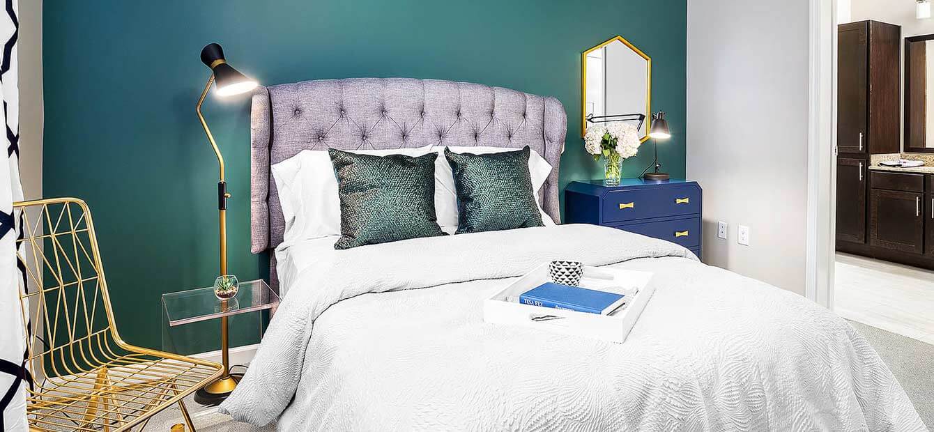 View of a bedroom. A lattice chair is on the left of the image. A large bed with a tufted headboard is in the center. A dresser is positioned next to the bed. A mirror is on the wall above the dresser. Next to the dresser is a door into a bathroom.